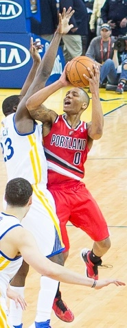 Damian Lillard 身穿拓荒者紅色客場球衣，比賽中在 Draymond Green 防守下騰空出手（授權 CC BY 2.0，來源 Wikimedia Commons）