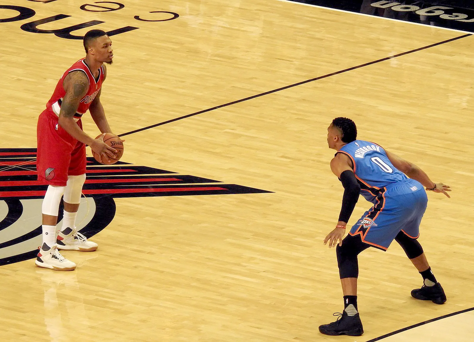 Damian Lillard 身穿拓荒者紅色球衣，比賽中持球面對 Russell Westbrook 的防守（攝影：James Schumacher，授權 CC BY 2.0，來源 Wikimedia Commons）