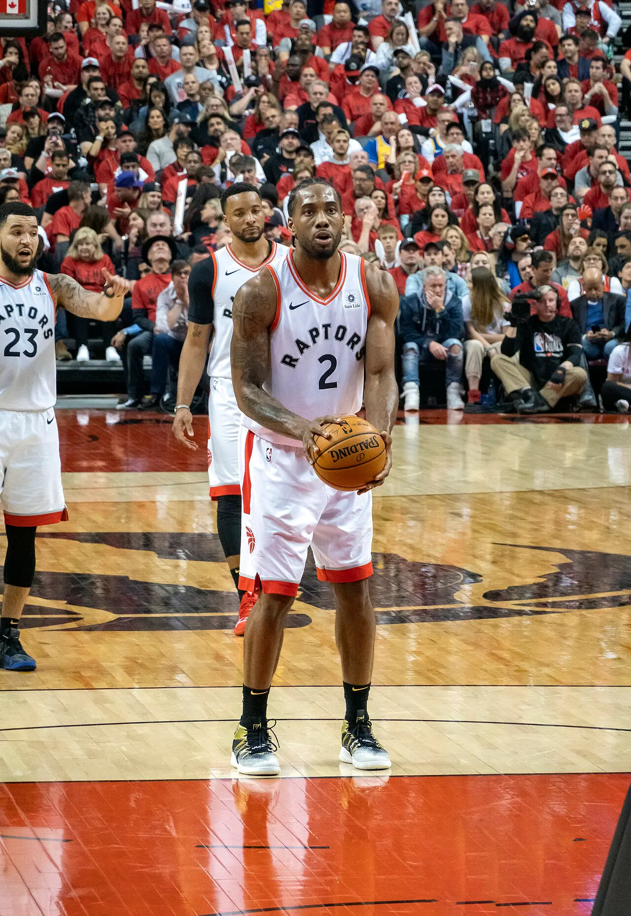 Kawhi Leonard 身穿暴龍白色 2 號球衣站上罰球線準備出手，左側為 23 號 Fred VanVleet，後方為滿場紅衣球迷（攝影 Chensiyuan，授權 CC BY-SA 4.0，來源 Wikimedia Commons）