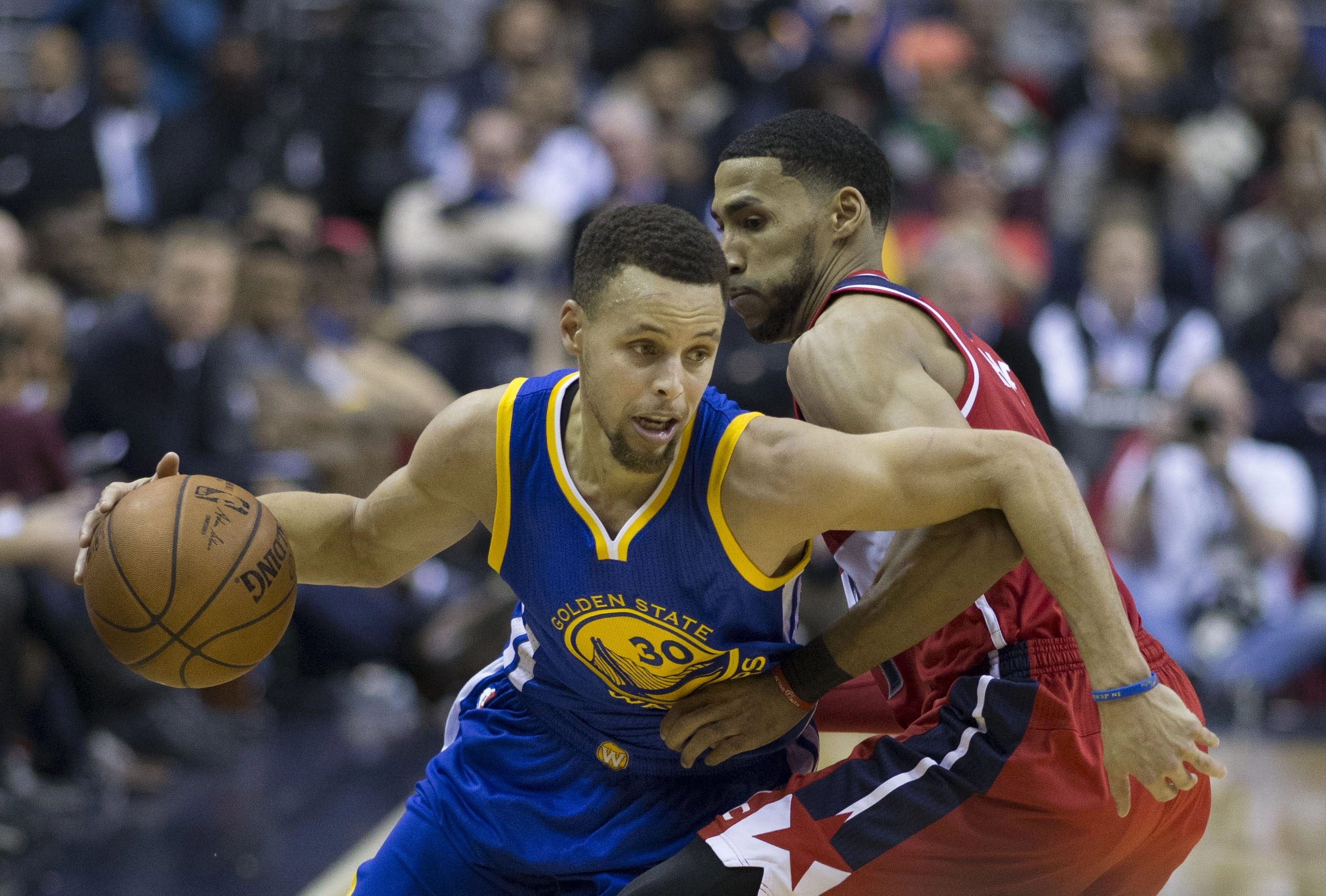 Stephen Curry 身穿勇士 30 號藍色球衣，比賽中護球突破，遭一名身穿紅色球衣的華盛頓巫師球員貼身防守。攝影：Keith Allison，授權 CC BY-SA 2.0，來源 Wikimedia Commons。