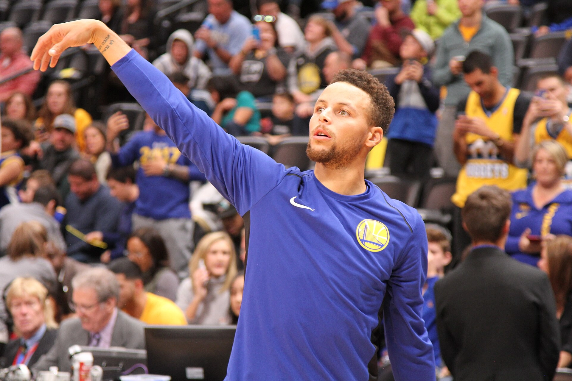 Stephen Curry 身穿勇士藍色長袖熱身衣，在丹佛客場賽前熱身時做出投籃出手後的跟隨動作。攝影：Cyrus Saatsaz，授權 CC BY-SA 4.0，來源 Wikimedia Commons。