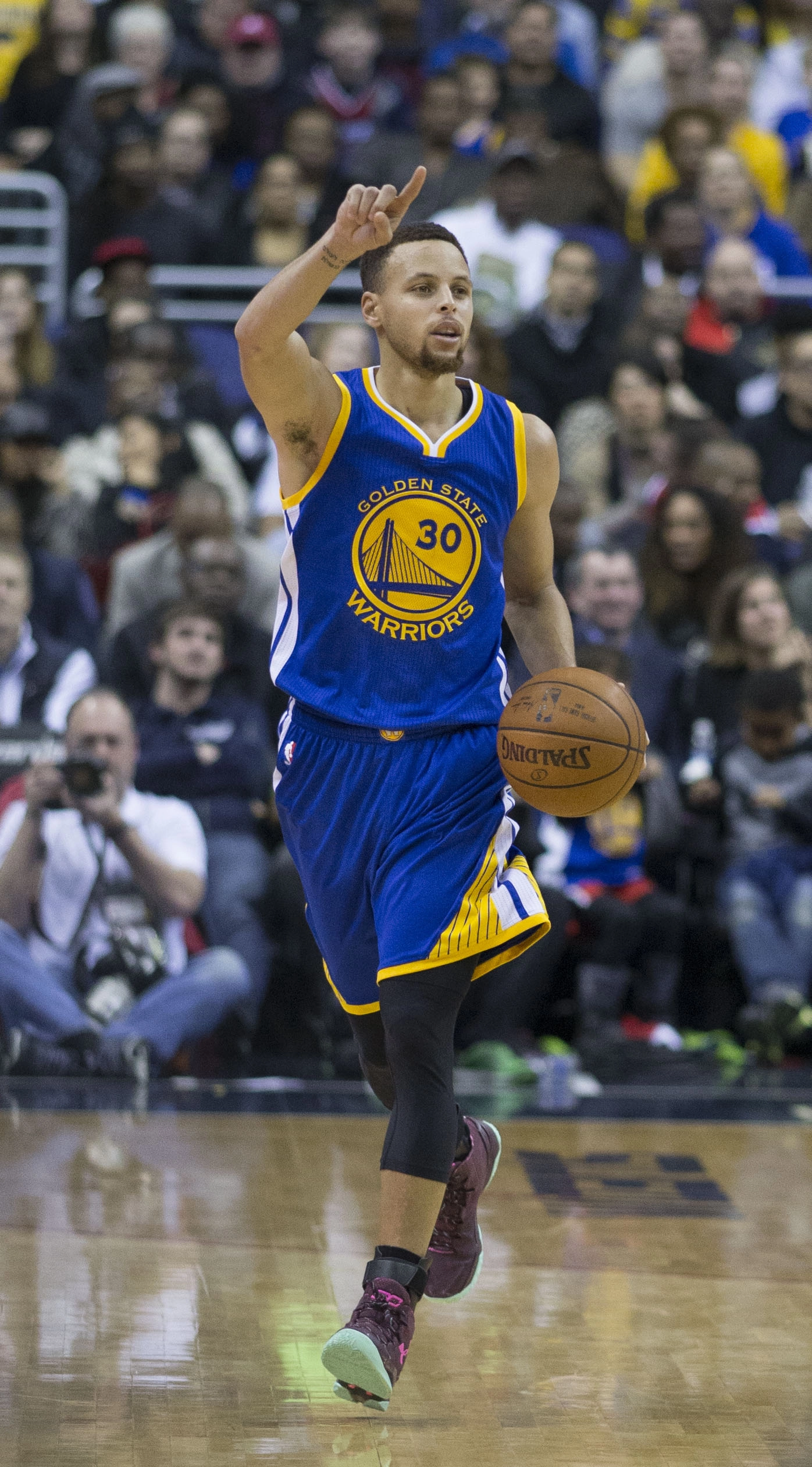 Stephen Curry 身穿金州勇士 30 號客場藍色球衣，比賽中運球推進並舉手指揮進攻。攝影：Keith Allison，授權 CC BY-SA 2.0，來源 Wikimedia Commons。
