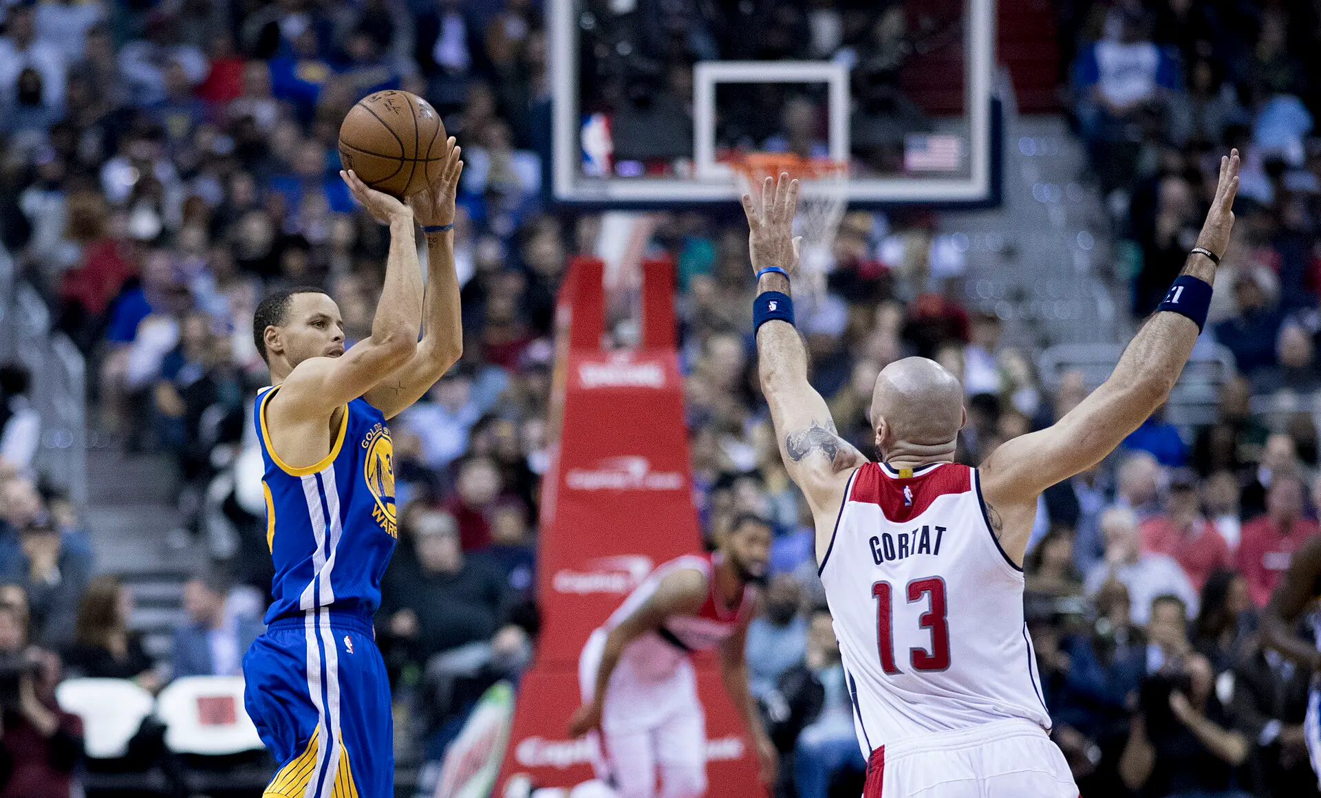 勇士 Stephen Curry 身穿藍色客場球衣跳投出手，巫師 13 號 Marcin Gortat 舉雙手防守（攝影 Keith Allison，授權 CC BY-SA 2.0，來源 Wikimedia Commons）
